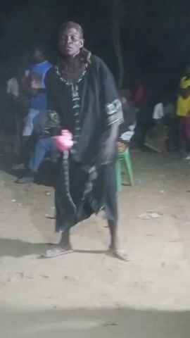 A single person, appearing to be a Black adult, is dancing in the center of a dirt lot. They are wearing a long black robe with embroidered cuffs and a small pink object is held in their hands. The dancer has red-tinted dreadlocks and wears sandals. They move around the lot, bending over and gyrating. A crowd of people is visible in the background, some seated, others standing. The scene is dimly lit.