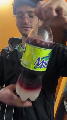 A young man with glasses and a black jacket is holding a partially filled plastic bottle of Manaos Pomelo Blanco soda, examining it closely. He holds the bottle upside down to get a better look at the contents. The bottle is clear with a purple-red liquid inside, and the label is bright green with purple lettering. The background is an orange wall, and the man has a black bag slung over his shoulder.