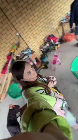 The video begins with a woman sitting on a chair, holding a vape and exhaling smoke. She is wearing a green hoodie with a cartoon character design and appears to be in an outdoor area with a brick wall in the background. The area is cluttered with various items, including shelves filled with objects, a small pink toy stroller, and some wooden planks. A person in shorts and a dark shirt is visible in the background, near a pile of items.

The scene then shifts to a blurry, fast-moving shot of an outdoor area with a pink table covered in items. Two red chairs are visible, one of which appears to be empty and the other partially obscured. In the background, there is a glimpse of a building with a green roof and a fence, suggesting a residential or rural setting. The overall impression is that of a casual, somewhat untidy outdoor space.

The time of day appears to be daytime, and the weather seems to be clear, as indicated by the lighting. The location context suggests that the video was filmed in or around Пепеляева, Russia. The woman's expression is neutral, and the activity appears to be casual vaping and filming. There is no visible text in the video.