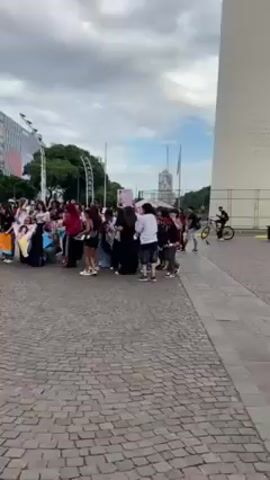 The video shows a group of approximately 30 people gathered in a public square, likely in a city. Most of the individuals appear to be young adults or teenagers. They are holding up signs with images, predominantly of a young man, possibly a celebrity or musician. Some signs have writing on them as well. The group is standing on a paved area with a cobblestone-like pattern. In the background, there is a tall, white obelisk-like structure, as well as a modern building with large signs. A person on a bicycle rides by in the distance. The weather appears overcast. The group seems to be expressing enthusiasm or support, possibly related to the person featured on their signs.