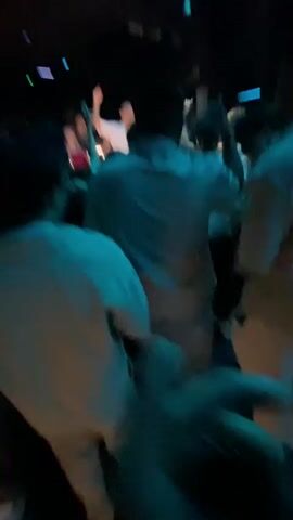 A video shows a crowd of people in a dimly lit room, likely a nightclub or concert venue. The lighting is predominantly blue, with neon accents and a spotlight directed at the center of the room where a person in a white shirt and hat appears to be performing or DJing. Several individuals are captured in motion, with hands raised, suggesting they are dancing or engaged with the performance. In the foreground, the backs of several people's heads are visible, further emphasizing the perspective of someone within the crowd. The ceiling features abstract neon designs.