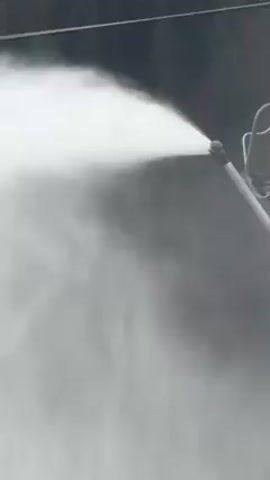 A video shows a snow cannon shooting a plume of white mist to the left with a ski lift in the background. The snow cannon is on the right side of the frame and angled to the left. The ski lift has two empty chairs hanging from it. Behind the ski lift and cannon is a forest that extends to the top of the frame. A power line runs horizontally across the frame at the top.