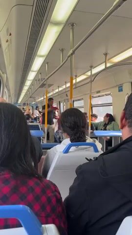 Here is a description of the video:

The video shows the interior of a train car. Several passengers are visible, seated and standing. One man stands in the aisle, holding a red cloth or item and speaking to other passengers. The seats are arranged in rows, and the train has poles for standing passengers to hold onto. There are windows on the side of the train and indicators above them. A restroom sign is visible near the end of the car. The lighting is bright and the overall scene appears to be ordinary.