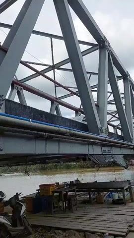 The video shows a view from a wooden dock looking up at a large metal truss bridge spanning a murky brown river. A white SUV is visible driving across the bridge, which also has pipes running along its underside. On the dock, there are several objects including a scooter, wooden crates, a table with stools, and buckets. The scene appears to be in a rural or semi-rural area with trees and vegetation visible on the far bank of the river. The weather is overcast with dramatic clouds, suggesting it might be daytime. The overall impression is one of a functional, perhaps somewhat utilitarian, transportation route in a tropical environment. There is no visible text in the video.