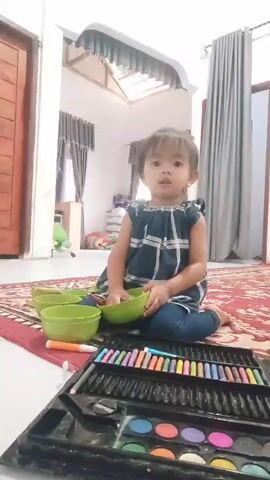 A young child with dark hair is sitting on a red rug in a brightly lit room. The child is wearing a dark blue dress with a white plaid pattern and blue pants. In front of the child are several green bowls and a box of crayons and watercolors. The child is holding one of the green bowls and appears to be playing with it. The room has light-colored walls, and in the background, there is a wooden door, gray curtains, and a skylight.