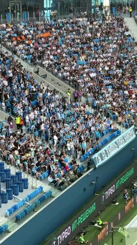 The video shows an aerial view of a crowded stadium filled with spectators. Most of the audience is wearing blue and white clothing, suggesting they are fans of a particular team. The stadium appears to be modern with tiered seating. In the foreground, there are blue seats and a green field with advertising banners. Some individuals are visible on the field, possibly staff or players. The overall atmosphere seems to be one of a sporting event or a large public gathering. The camera is positioned high above, looking down at the scene.