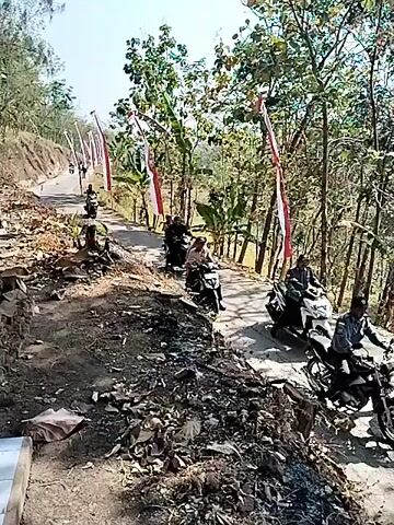 A group of people on motorcycles are traveling along a dirt road. The road is lined with trees, and there are Indonesian flags on the side of the road. The people appear to be participating in a procession or parade. The weather is sunny.
