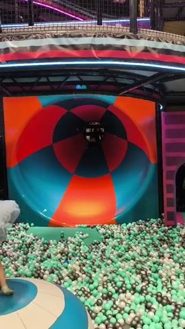 A child slides down a large, colorful spiral slide into a ball pit. The slide is red, orange, and teal, with a circular pattern. The ball pit is filled with green, white, and black balls. Two other children are visible in the foreground, one on the left and one on the right, watching the child slide. The background shows a play structure with netting and neon lights.