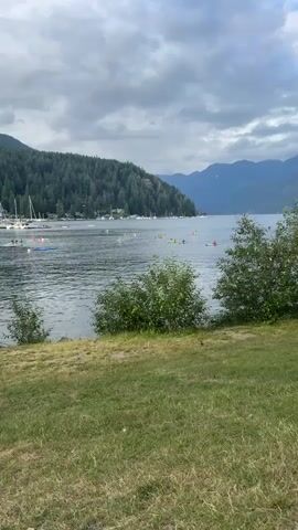 The video captures a serene waterscape with a lush green grassy foreground. In the distance, a calm body of water is dotted with numerous kayaks and some sailboats, suggesting a recreational activity. The water is surrounded by a dense forest that blankets the rolling hills, with mountains forming a majestic backdrop under a cloudy sky. A small bird is seen foraging on the grass in the foreground, adding a touch of wildlife to the peaceful scene.