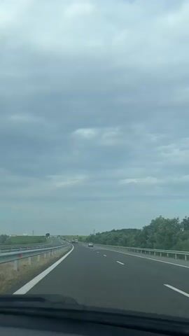 A point-of-view shot from inside a moving vehicle, looking forward and to the side, shows a highway. The road is paved with asphalt and has white lane markings. A metal guardrail runs along the side of the road, separating it from a grassy area and trees. The sky is cloudy, with patches of blue visible. There are a few other vehicles on the highway in the distance. The interior of the vehicle is visible in the foreground, including part of the dashboard and windshield. A side mirror is also visible on the right side of the frame. The trees are lush and green, suggesting a temperate climate. The overall impression is of a journey on a highway under a partly cloudy sky.