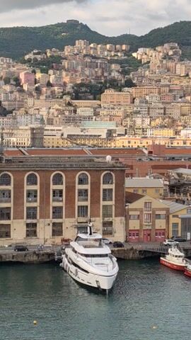 A long shot shows a city next to the water. Buildings are close together and stretch up the side of a hill. On the other side of the city are several boats docked. A long, thin mast extends into the sky.