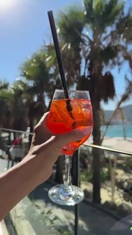 A close-up shot shows a hand holding a glass of an orange beverage with ice and an orange slice. The beverage is in a stemmed wine glass with a black straw. In the background, a row of palm trees lines a beach with white umbrellas and lounge chairs. The ocean and a distant island are visible under a clear blue sky. The camera pans slightly to the right, revealing more of the beach scene.