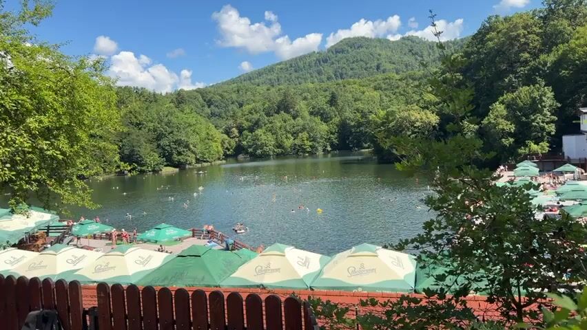 The video shows a wide shot of a lake surrounded by lush green trees and a mountain in the background. The sky is blue with scattered white clouds. On the left side of the lake, there is a beach area with many people swimming and sunbathing. There are also several green and white umbrellas providing shade. On the right side of the lake, there are more umbrellas and some buildings. The water is calm and reflects the trees and sky. Several people are visible in the water, some swimming and others floating on inflatable devices. The overall impression is a lively and popular summer resort.