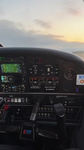 The video shows the interior of a small airplane cockpit. The camera is positioned behind the pilot, looking forward towards the dashboard and out the windshield. The dashboard is filled with various instruments and controls, including screens displaying flight information. The pilot's hands are visible on the yoke. The view through the windshield shows a runway ahead, with a cloudy sky and the sun setting in the distance. The plane appears to be on the runway, possibly preparing for takeoff or landing. The overall impression is one of pre-flight or post-flight activity in a light aircraft.