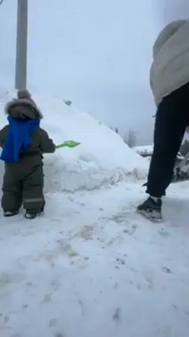 The video shows a person shoveling snow next to a small child in a snowy outdoor environment. The child, wearing a green snowsuit, a blue scarf, and a knitted hat with a pom-pom, holds a small green toy shovel. The person, dressed in a white puffy jacket and black pants, is using a black snow shovel to clear the area near a large snow pile. A utility pole is visible to the left, and a green building is partially visible in the background. The ground is covered in snow, and the sky is overcast.