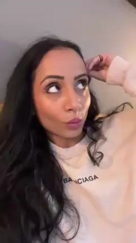 A woman with long, dark, wavy hair and pink lipstick is smiling at the camera. She is wearing a light-colored sweatshirt with "Banciaga" printed on it. Her left hand is near her forehead, as if she's resting it there. The background is a blurry, neutral color, possibly a wall. The lighting suggests an indoor setting. She is the sole subject in the frame.