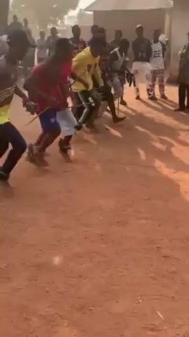 A group of young men, mostly wearing colorful sports clothes, are dancing in a line on a dirt ground. They are bending over in unison and kicking their legs out, stirring up dust. Some are holding leafy branches. A crowd of onlookers, also mostly men, surrounds them. There are buildings in the background.