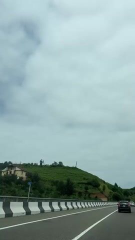 The video shows a view from a car driving on a highway. On the left side of the road is a grassy hill with some trees and a house. On the right side of the road, other cars are visible. The sky is cloudy.