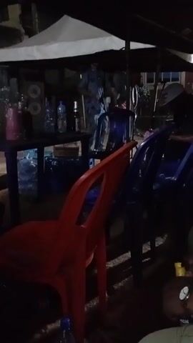 The video shows an outdoor gathering at night, under a white canopy. There is a dark table with water bottles and a beer bottle on it, next to a box. Several plastic chairs, in orange and blue, are arranged in front of the table. People are walking and standing behind the table, illuminated by the light. One person is wearing a red shirt. The area is dimly lit and the focus is on the activity under the canopy. Music is playing in the background.