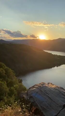 The video shows a sweeping drone shot over a large body of water surrounded by rolling hills and mountains. The sun is setting, casting a warm golden light on the landscape and creating a serene and picturesque scene. The water is calm and reflects the sky and surrounding scenery. The hills are covered in lush green vegetation, with some areas appearing to be wooded. The mountains in the distance are silhouetted against the sunset sky. In the foreground, a large rock formation is visible, with some dry grass and small flowers growing around it. The drone moves smoothly, offering a panoramic view of the natural beauty. There are no people or man-made structures visible in the video, emphasizing the untouched and wild nature of the environment.
