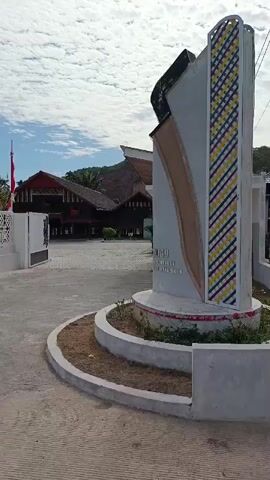 A medium-shot, slightly angled video begins by showing a tall, modern monument on the right side of the frame. The monument is white with geometric, colorful patterns on its right side, and a brown accent on its left. The monument is situated on a circular concrete base with a brown, mulched area inside. In the background, to the left of the monument, is a courtyard with a paved surface. Further in the background, there are several traditional-looking buildings with dark wood and steep, dark roofs. In front of the buildings, there are several flagpoles flying red and white flags. A white, ornate fence runs along the left side of the courtyard. A person in red clothing is visible crouching in the lower left corner of the frame. The sky is cloudy with white clouds against a light blue background. The video pans slightly to the left, revealing more of the courtyard and buildings.
