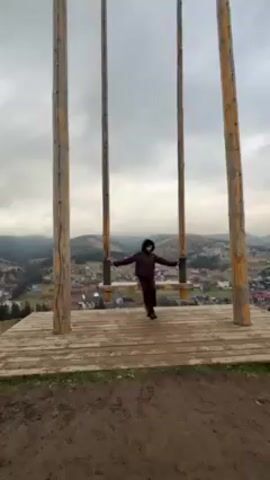 The video shows a young woman sitting on a swing set overlooking a small town in the mountains. The woman is wearing a dark brown puffer jacket and pants and a black beanie. The swing set is made of wood and has four large wooden posts. The platform the swing sits on is also wooden. The town below has many houses and buildings, and the mountains are covered in trees. The sky is cloudy and overcast.