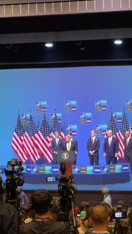 A group of men stand behind a podium in front of a blue backdrop with the NATO logo and the word "ANKARA SUMMIT". Three American flags are positioned to the left of the podium. A man in a dark suit and yellow tie, identified as Donald Trump, is speaking at the podium. Two other men in suits are standing to his left and right. The audience is visible in the foreground, with cameras and microphones set up.