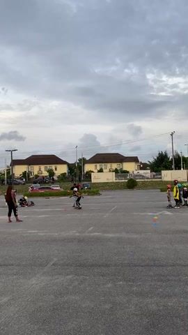 The video shows a group of people in a parking lot. There are several children learning to rollerblade, under the supervision of adults. Orange cones are placed on the ground, which the children are skating around. The parking lot is empty except for a few parked cars. Two yellow houses are in the background. The sky is cloudy. One adult is standing in the middle of the parking lot and seems to be instructing one of the children. Another adult is standing further away, observing the group.