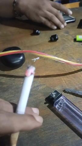 A close-up shot shows a person's hand holding a lit cigarette, with the cherry glowing. In the background, on a wooden table, are several objects: a clear plastic lighter, a serrated knife, a pair of black earbuds, a black oval-shaped stone, and a bundle of yellow and red wires. Another person's hands are visible in the upper portion of the frame, resting on a smartphone. The scene appears to be indoors, possibly a casual setting. The lighting suggests it might be daytime, but the specific time of day is not discernible from the image. No discernible emotions are conveyed. The visible text is limited to what appears to be a brand name on the lighter, though it is not clearly legible. The location context is Keudematangkuli, Indonesia.