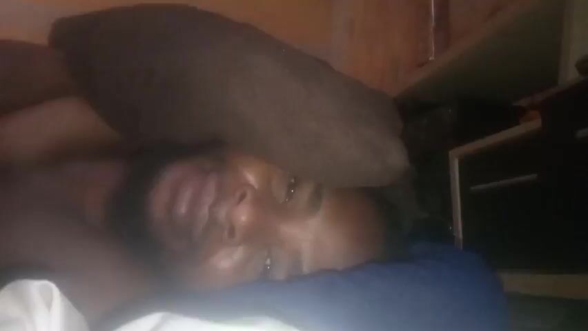 A person is lying down, possibly in bed, with their head on a blue pillow. They are holding up their hand in front of their face, obscuring part of their vision. A brown pillow is behind their head. A nightstand with a drawer is visible in the background. The lighting in the room is dim.