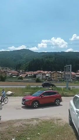 This media file captures a sunny daytime scene in Yakoruda, Bulgaria. The camera is positioned at a slight upward angle, looking towards a cluster of houses nestled at the foot of a lush, tree-covered mountain range under a bright blue sky dotted with fluffy white clouds. 

In the foreground, several vehicles are visible. A white SUV is parked closest to the camera, its windshield reflecting the sunlight. To its left, a red car is also parked. Further back, a dark-colored SUV is driving on a road that runs horizontally across the middle ground.

The houses in the village appear to have red-tiled roofs, a common architectural style in many European regions. The mountain slopes are densely forested, suggesting a rural or semi-rural environment.

The overall atmosphere is peaceful and summery. The clear sky and bright sunlight indicate fair weather. The presence of the village and the surrounding natural landscape suggests a typical day in a Bulgarian town. There are no discernible signs with visible text in this clip. The clip appears to be a short, unedited segment of everyday life.