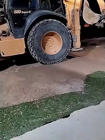 The video shows a Case 580 Super R backhoe parked at night near the edge of a concrete sidewalk and a patch of grass. The backhoe is illuminated by its own headlights and possibly other unseen light sources. The backhoe's left rear tire is prominently featured, along with the machine's yellow-painted backhoe arm and bucket, which are raised in the air. Beyond the backhoe, the background fades into darkness, with faint lights visible in the distance. There are also miscellaneous items visible on the ground. The camera is mostly stationary.