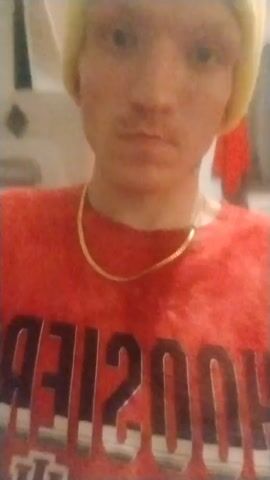 A person wearing a light-yellow beanie and a red t-shirt with "HOOSIERS" printed in reverse with the Indiana University logo on it is facing the camera. The person also has a gold chain necklace. The background appears to be a bathroom.
