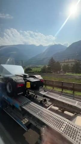 The video shows a truck with the number 380 on its cab, identified as HVO, transporting another truck on its flatbed. The trucks are on a highway with mountains in the background. The sun is shining brightly, and there are some clouds in the sky. A train track runs parallel to the highway, with a wooden fence separating them. The overall scene depicts a sunny day in a mountainous region, with trucks on a road and a train track.