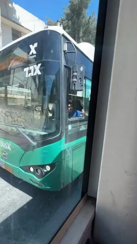 The video shows the front of a green and white bus with the "X" logo and Hebrew text on the windshield. A man, presumably the driver, is visible in the driver's seat, looking out the window and waving. The bus appears to be stopped on a street. The scene is brightly lit, suggesting it is daytime with clear weather. In the background, there are buildings and trees. The text on the bus, in Hebrew, includes "X" and "TAV," possibly indicating a bus line or company. The reflection in the bus windshield suggests the video is being filmed from inside another vehicle, possibly another bus or car, looking out a window. The interior of the vehicle from which the video is filmed is partially visible on the right side of the frame, showing a white surface and part of a seat. The overall atmosphere appears casual, with the driver acknowledging the camera.