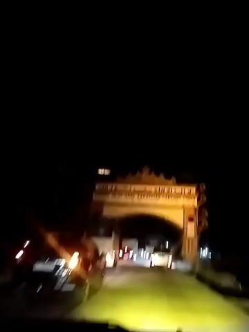 A nighttime video shows a car driving through an archway illuminated by lights. The archway has writing on it in Arabic script and what appears to be English. Traffic, including trucks and cars, is visible on the road under and beyond the archway. The scene suggests the car is passing through a border crossing or an entrance to a town or region.