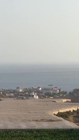 The video shows a wide shot of a coastal landscape. In the foreground, there are green plants, possibly crops, with a textured surface in front of them. The middle ground features a low-lying town or resort with white buildings and red-tiled roofs, situated near the coast. A body of water, likely the ocean, extends into the background, with a hazy horizon. A small boat is visible on the water in the distance. The sky is bright and overcast. There are no people or specific actions depicted. The overall impression is a scenic view of a coastal settlement against the backdrop of the sea and sky.
