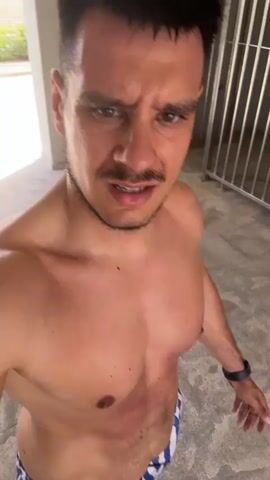 A shirtless man with short dark hair and a mustache is looking at the camera. He is in what appears to be an outdoor or semi-outdoor space with a concrete ceiling and a metal railing in the background. He is wearing blue and white striped shorts. The man has a concerned or stressed expression on his face. The lighting suggests it is daytime.