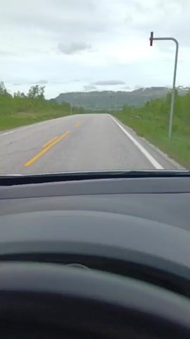 The video shows the view from inside a car driving on a paved road. The road has a double yellow line in the center and a white line on the right edge. The landscape on either side of the road is green and hilly, with some trees. In the distance, there are mountains under a cloudy sky. The dashboard and steering wheel of the car are visible in the foreground. The car appears to be moving at a moderate speed.