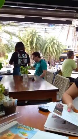 The video shows the interior of an open-air cafe or restaurant, presumably in a warm, tropical location due to the presence of palm trees outside the windows. The cafe has multiple tables, some occupied by patrons. 

Several individuals are seated at different tables. A woman in the foreground is holding a menu and looking at it. The camera pans around to show other diners sitting at their tables, including one group where people are also looking at menus.

The cafe has a modern, somewhat minimalist design with wooden tables and woven chairs. The ceiling is dark and has a grid-like structure. Bright daylight filters through the open walls, illuminating the interior.