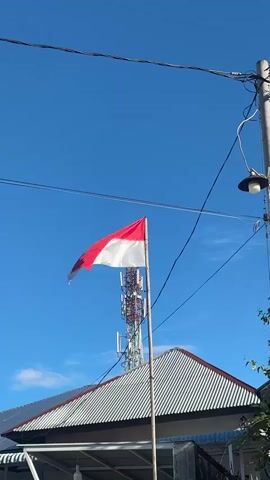 A low-angle, outdoor, vertical video shows the Indonesian flag flying in the wind against a clear blue sky. The flag, which is red on top and white on the bottom, is attached to a metal pole. Behind the flag, a tall telecommunications tower with multiple platforms and antennas is visible. In the foreground, the top of a building with a corrugated metal roof is partially visible, along with overhead power lines and a street lamp. The scene appears to be in a populated area, possibly a town or city.