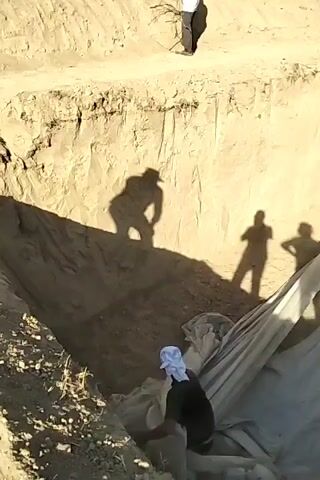 The video shows a steep excavation with several people present. In the foreground, a person wearing a white head covering and dark clothing is kneeling and appears to be working with a large sheet of white fabric. In the middle ground, several large shadows of people are cast on the side of the excavation. One shadow is of a person wearing a hat, bending over. Two other shadows are of people standing. In the background, a person is climbing a ladder out of the excavation. The excavation is dug into dry, sandy soil, and the fabric appears to be laid out or spread within it. The overall scene suggests construction or some form of earthmoving activity.