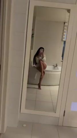 A woman with long black hair is sitting on the edge of a white bathtub in a white bathroom, taking a photo of herself in a full-length mirror. She is wearing a white, loose-fitting shirt or robe and white bottoms. The bathroom features white tiled walls and floor. A white towel hangs next to the bathtub. There is a window with blinds in the background. The woman is holding a white phone, presumably taking the picture.