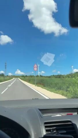 The video is filmed from the perspective of someone in the passenger seat of a car, looking out the windshield. The car is driving on a paved road that curves to the left. On the right side of the road, there is a grassy field with red flowers and a few wind turbines. In the distance, there is a power tower and more wind turbines. The sky is bright blue with some white clouds. On the windshield, there are two stickers. One is a white square with some text and a QR code. The other is a circular sticker with a pattern of dots. Hanging from the rearview mirror are two black dice on a chain.