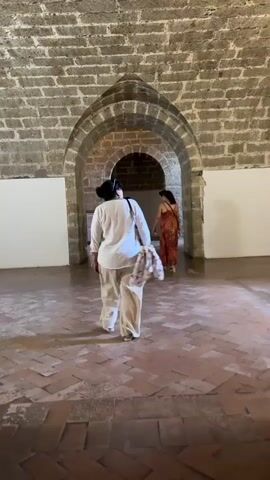 Two women are walking through a stone hallway. The woman in the foreground is wearing a white shirt and pants, and the woman in the background is wearing a patterned dress. The hallway is made of stone and has arched doorways. The floor is paved with bricks.