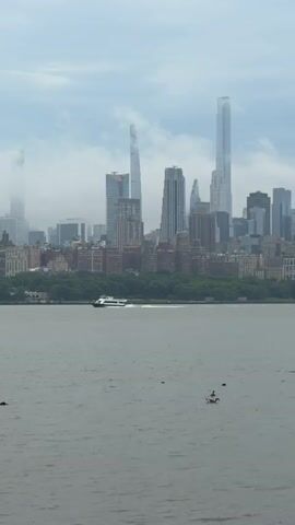 This media file captures a panoramic view of the New York City skyline from across the Hudson River. The skyline is dominated by a cluster of skyscrapers, including several extremely tall, slender buildings, some of which are partially obscured by a dense, gray, overcast sky. The weather appears to be cloudy and possibly humid, contributing to a muted color palette.

In the foreground, the murky, gray waters of the Hudson River stretch across the frame. A white ferry boat is seen moving from left to right across the river, leaving a white wake behind it. On the water, several birds, likely ducks or geese, are visible. One pair is swimming together, while another bird is further to the left. The shoreline opposite the viewpoint is lined with a strip of green trees, indicating a park or natural area along the edge of the city. Beyond the trees, the mid-rise buildings of the city form a dense urban landscape that gives way to the towering skyscrapers.

The overall mood is somewhat somber due to the overcast weather, but the presence of the moving ferry and the birds adds a sense of life and activity to the scene. The time of day is not explicitly clear, but the diffused light suggests it could be late morning or afternoon. The location is confirmed as North Bergen, United States of America, indicating the viewpoint is from the New Jersey side of the Hudson River, looking towards Manhattan. There is no visible text in the media file.