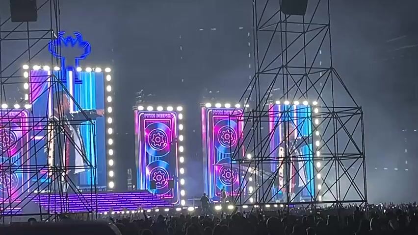 A large concert stage is lit with bright lights and screens displaying colorful graphics. The stage is surrounded by scaffolding. A crowd of people is visible in the foreground, appearing to be watching the performance. A person is standing on the stage, facing the crowd. The lighting is dynamic, with flashing lights and spotlights. The overall atmosphere suggests a lively and energetic event.