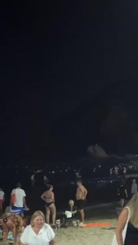 The video captures a nighttime scene on a beach in La Cala, Spain. The primary subjects are groups of people on the sand and in the water. In the foreground, several individuals are visible, including two women talking, one wearing a black bikini and the other a white top, and a man in swim trunks. Further back, many people are in the ocean, appearing to be swimming or socializing. A prominent object in the foreground is a skeleton on a chair, which seems out of place.

The activity taking place is a beach gathering or party at night. The presence of many people in the water and on the shore, combined with the dark sky, suggests a lively, informal event. The time of day is clearly night, indicated by the darkness and the presence of artificial lights in the distance, likely from the town. The sky is dark, with no visible clouds, and a full moon is visible. The ocean appears to have moderate waves.

Notable details include the illuminated skeleton figure, which adds a quirky or perhaps festive element to the scene. The overall mood appears relaxed and social, with people enjoying the beach after dark. The location cues, like the mention of La Cala, Spain, help establish the setting. There is no visible text in the media. The dark, somewhat grainy quality of the video is consistent with low-light nighttime recording.