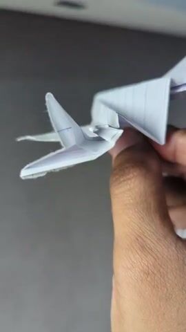 A close-up shot shows a hand holding a white paper airplane against a gray textured background. The airplane has a pointed nose, swept-back wings, and a vertical stabilizer. The video loops, showing the airplane from slightly different angles in each frame.