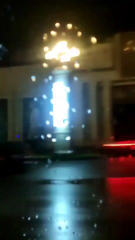 The video captures a night-time drive-through scene. The camera is inside a vehicle, looking out. The primary focus is on buildings passing by, illuminated by streetlights and possibly interior lights. Some of the buildings appear to be businesses, with large windows and architectural features like columns. There is a visible tree planted outside one of the buildings. The road surface is dark and wet, suggesting rain. Reflections and water droplets are visible on the vehicle's window, indicating the presence of rain or moisture. A dark-colored vehicle, possibly an SUV, is visible in the foreground, with its taillights illuminated. The overall atmosphere is dark and blurry due to the motion and low light conditions.